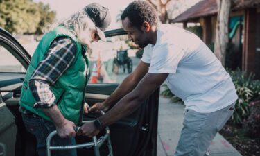 A man helps another man with a walker get out of his car.