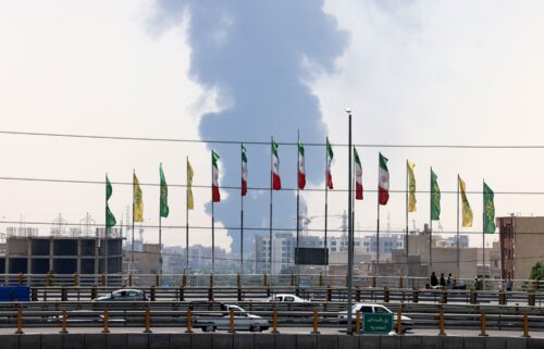 Flags flutter along a bridge as a plume of heavy smoke and fire rises from an oil refinery in southern Tehran on June 15